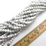 Flera trådar av Howlit rund ädelstenpärlor, vita med grå marmorering och ca 6mm i diameter, är arrangerade vid en trälinjal.