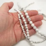 En hand håller i trådar av Howlit rund ädelsten 4-8 mm runda vita pärlor med grå marmorering mot en vit bakgrund.