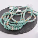 En lång tråd av Amazonit flerfärgad rund smyckesten 6mm pärlor i ljusgrönt, vitt, solbrunt och ljusblått är upprullad på en mörk skifferplatta med flera vita taggar.
