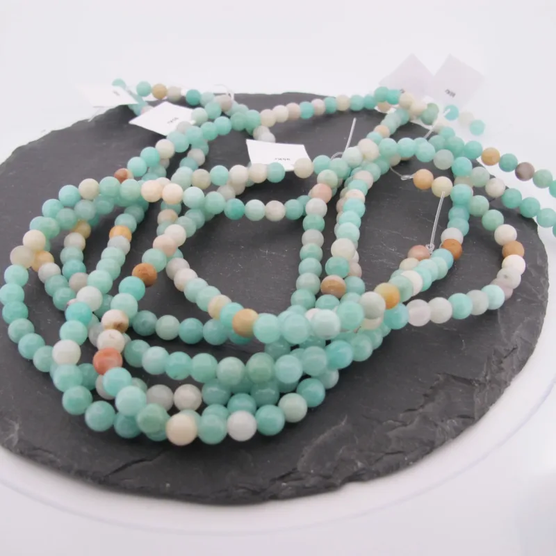 En lång tråd av Amazonit flerfärgad rund smyckesten 6mm pärlor i ljusgrönt, vitt, solbrunt och ljusblått är upprullad på en mörk skifferplatta med flera vita taggar.