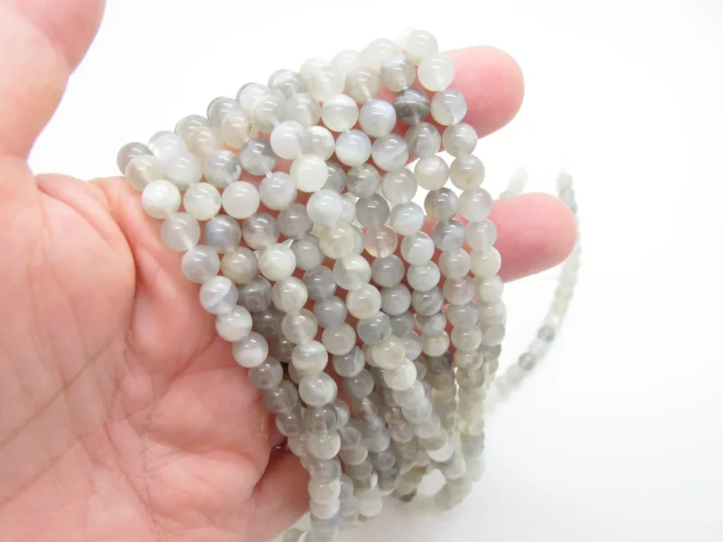 En hand håller i flera trådar av Månsten rund ädelsten 6mm, med runda, halvgenomskinliga grå och vita pärlor mot en enfärgad vit bakgrund.