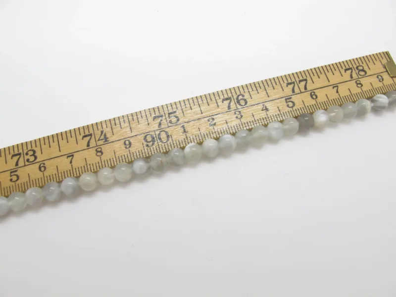 En Månsten rund ädelsten 6mm pärlsträng - en rad med små, runda, ljusa pärlor - mäts med en trälinjal som är märkt i tum och centimeter.