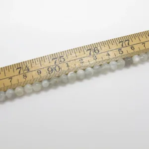 En Månsten rund ädelsten 6mm pärlsträng - en rad med små, runda, ljusa pärlor - mäts med en trälinjal som är märkt i tum och centimeter.