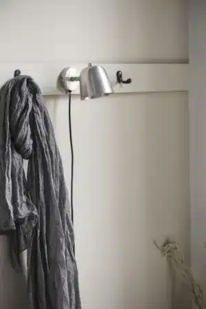 En ljusgrå scarf hänger från en Krok dubbel mini - mässing krok på en benvit väggmonterad hylla. I närheten hänger en silverfärgad vägglampa med svart sladd och en annan tom mässingskrok är synlig. Bakgrundsväggen är offwhite.