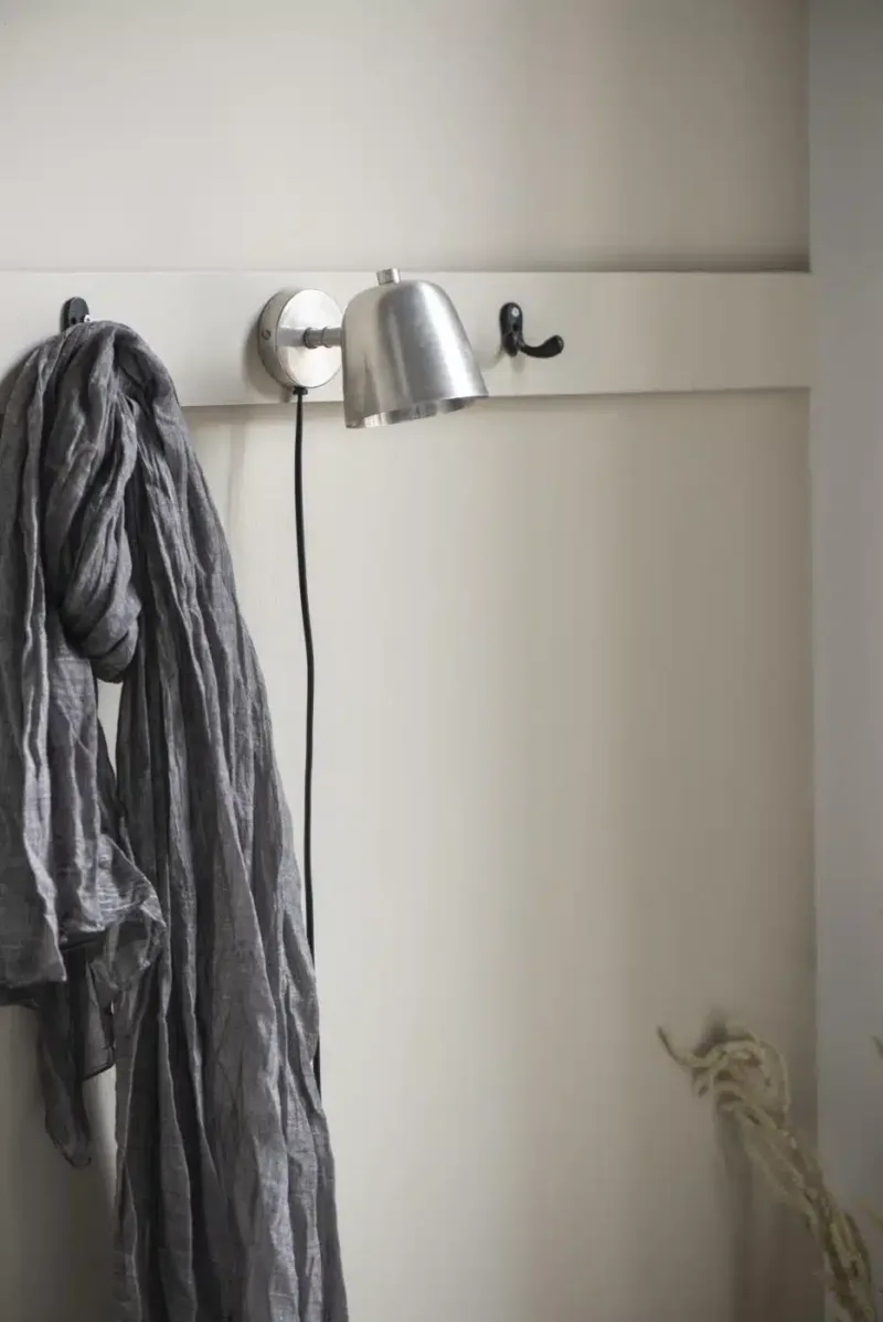 En ljusgrå scarf hänger från en Krok dubbel mini - mässing krok på en benvit väggmonterad hylla. I närheten hänger en silverfärgad vägglampa med svart sladd och en annan tom mässingskrok är synlig. Bakgrundsväggen är offwhite.