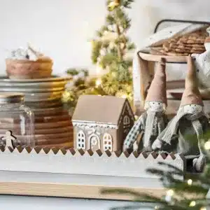 En mysig julscen med Pepparkakshus för ljus med dörrkrans pepparkakshus, två tomtedockor i tyg, en dekorerad minigran, staplade tallrikar, kakor och varma ljusslingor på bordet.