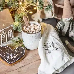 En mysig scen med Pepparkakshus för ljus med dörrkrans, en mugg varm choklad med marshmallows, en hjärtformad pepparkaka, en tänd minitall, en servett och en tomtedocka i tyg på en träyta.