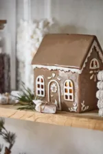 Pepparkakshuset för ljus med dörrkrans, ett keramikhus med glasyrdetaljer, lyser varmt på en hylla bredvid inslagna godisbitar, tallkvistar och burkar med marshmallows och choklad.