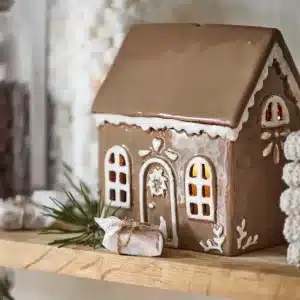 Pepparkakshuset för ljus med dörrkrans, ett keramikhus med glasyrdetaljer, lyser varmt på en hylla bredvid inslagna godisbitar, tallkvistar och burkar med marshmallows och choklad.