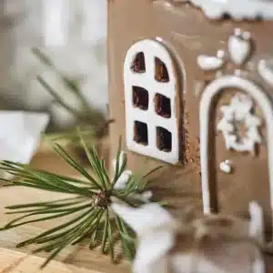 Närbild på keramikhuset Pepparkakshus för ljus med dörrkrans, med vita glasyrdetaljer, fönster och dörrkrans, prytt med tallgrenar och en liten inslagna present på en träyta.