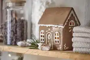 Pepparkakshus för ljus med dörrkrans, ett pepparkakshus i keramik, vilar på en trähylla bredvid en glasburk med choklad, små inslagna presenter, tallkvistar och hopvikta stickade dukar.