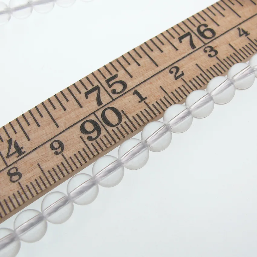 Bergkristall 6mm rund ädelsten 3 Close-up of a wooden measuring tape showing 74–76 cm with a string of translucent white beads laid diagonally across it.