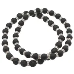 Lavasten svarta runda 4-8mm vaxade halsband har omväxlande matta och texturerade svarta lavapärlor, åtskilda av silverfärgade distansringar med små klara kristaller. Halsbandet visas här upprullat i en lös spiral.