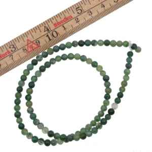 Ett pärlband med Mossagat rund ädelstenspärlor (4-8mm) i olika gröna nyanser är arrangerat i en ögla bredvid en trälinjal markerad i centimeter. Stenpärlorna varierar i färg och storlek.