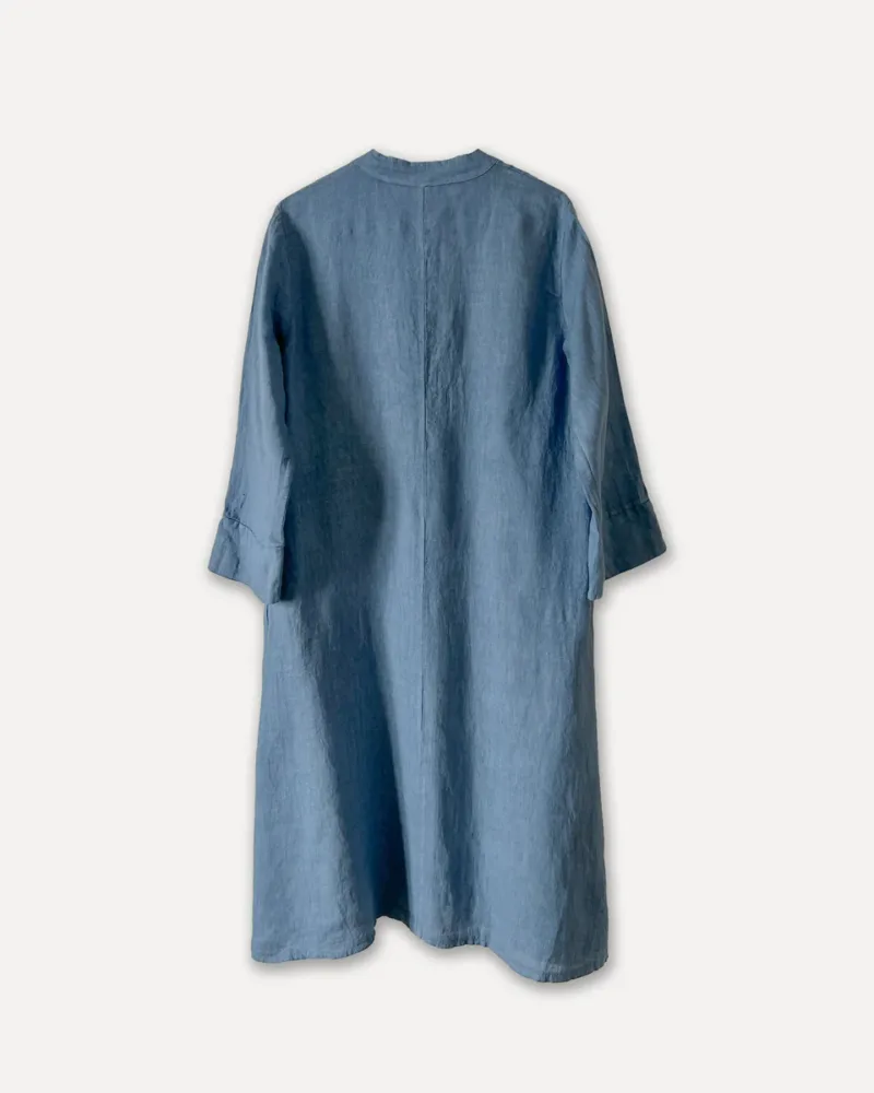 En löst sittande, långärmad, ljusblå linneklänning med enkelt mönster, visad bakifrån mot en enfärgad vit bakgrund.