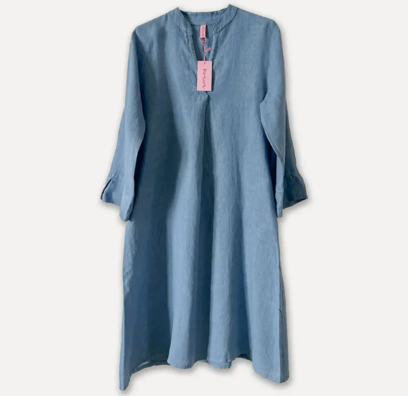 En långärmad, ljusblå linneklänning med enkel krage och en rosa tagg fäst nära halsringningen, mot en vit bakgrund.