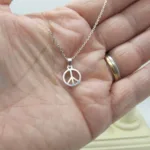 En hand håller i ett silverhalsband med ett litet runt hänge med en fredssymbol. Personen bär två ringar på ringfingret, en i guld och en i silver.