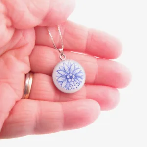 En hand håller i ett runt hänge på en silverkedja med en blå blomsterdesign mot en vit bakgrund. Personen bär en guldring på sitt finger.