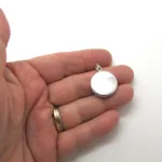 En hand håller i en liten, rund silvermedaljong mot en vit bakgrund. Personen bär en vigselring i guld.