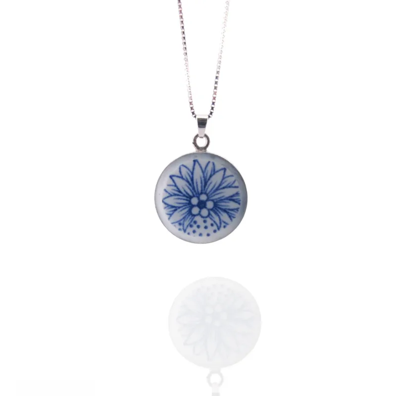 Porslinsmycke - Dresden 29x19mm är ett silverhalsband med ett runt hänge med ett blått handmålat blomstermotiv på en vit bakgrund, som visas tillsammans med sin reflektion på en vit yta.