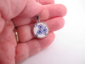 En hand håller i ett runt halsband med en vit bakgrund med en blå blommig design, infattat i en metallram.