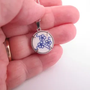 En hand håller i ett runt halsband med en vit bakgrund med en blå blommig design, infattat i en metallram.