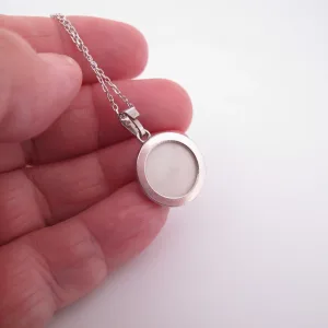 En hand håller i ett silverhalsband med en rund, vit mitt, möjligen en medaljong eller en minnessak, mot en vit bakgrund.