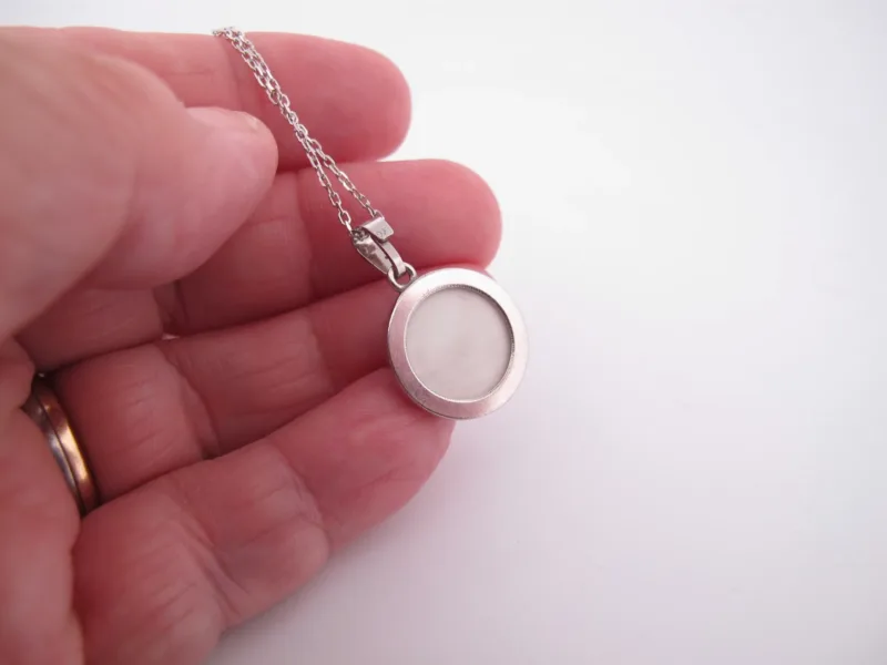 En hand håller i ett silverhalsband med en rund, vit mitt, möjligen en medaljong eller en minnessak, mot en vit bakgrund.