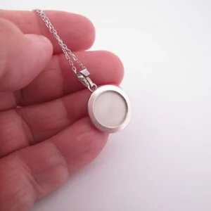 En hand håller i ett silverhalsband med en rund, vit mitt, möjligen en medaljong eller en minnessak, mot en vit bakgrund.