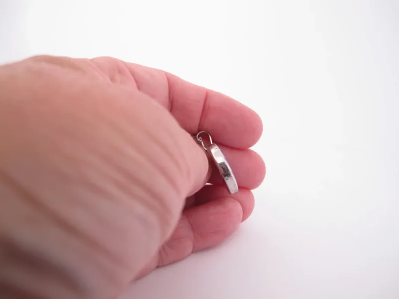 En hand håller i ett litet ringformat silverhänge mot en vit bakgrund.