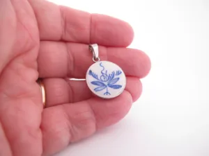 En hand håller i ett runt hänge med blått blommönster mot en vit bakgrund, med en ögla i silvermetall för att fästa på ett halsband.