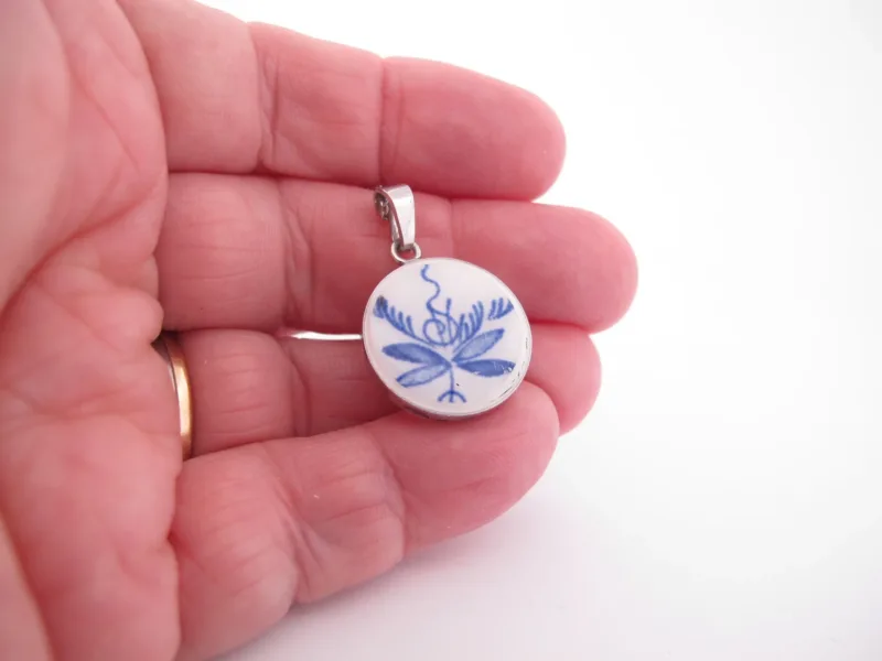 En hand håller i ett runt hänge med blått blommönster mot en vit bakgrund, med en ögla i silvermetall för att fästa på ett halsband.