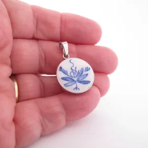 En hand håller i ett runt hänge med blått blommönster mot en vit bakgrund, med en ögla i silvermetall för att fästa på ett halsband.