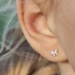 Närbild av en persons öra som bär ett litet fjärilsformat örhänge i silver. Bakgrunden är mjukt suddig, vilket framhäver örhänget och en del av personens hår.