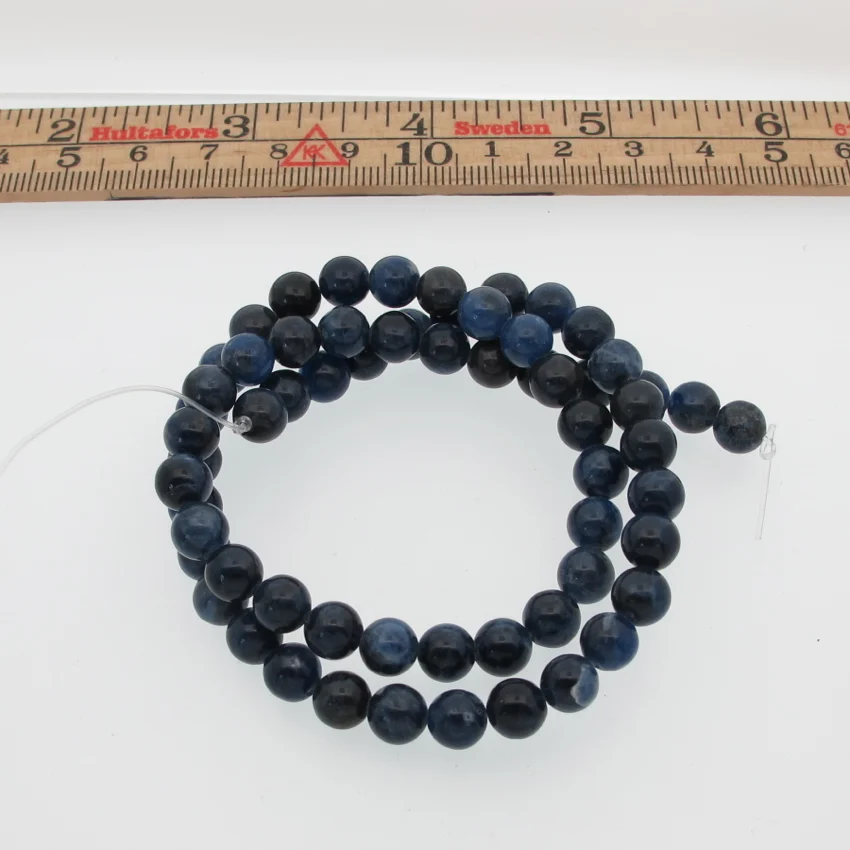 Sodalit 6mm rund ädelsten 2 En spiralformad tråd av Sodalit runda ädelstenspärlor (4-6 mm i diameter) ligger på en vit yta under en linjal och framhäver deras runda, mörkblå utseende.