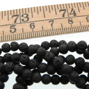 Närbild av en trälinjal ovanför flera strängar av Lavasten svarta runda 4-8mm vaxade, små runda svarta vaxade lavapärlor, arrangerade på en vit yta.