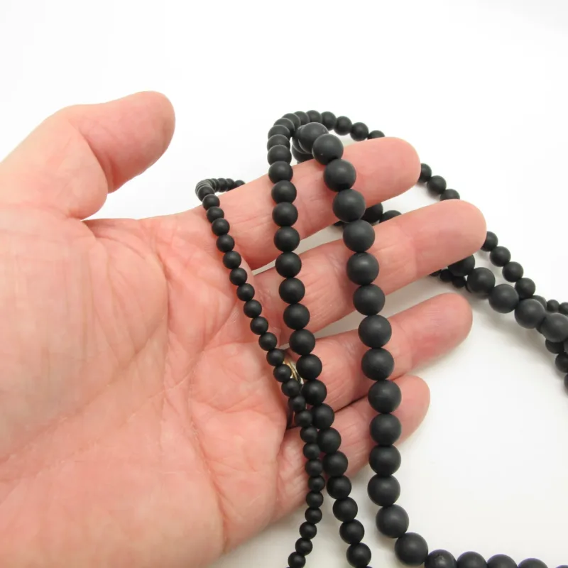 En hand håller i en tråd av Mattsvart Onyx rund ädelstenspärlor (4-10mm) mot en vit bakgrund.