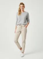 En kvinna står mot en enfärgad bakgrund och ler. Hon är klädd i en ljusgrå tröja, beigea byxor med dragsko och vita sneakers, med håret uppsatt i hästsvans. Hon har ena handen i fickan.