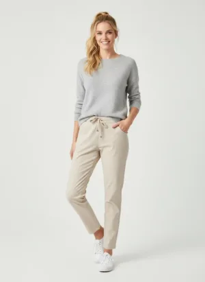 En kvinna står mot en enfärgad bakgrund och ler. Hon är klädd i en ljusgrå tröja, beigea byxor med dragsko och vita sneakers, med håret uppsatt i hästsvans. Hon har ena handen i fickan.