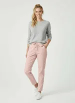 En kvinna står leende med ena handen i fickan. Hon bär en ljusgrå tröja, ljusrosa byxor med dragsko i midjan och vita sneakers. Hennes blonda hår är uppsatt i en lös hästsvans. Bakgrunden är enfärgad och ljus.