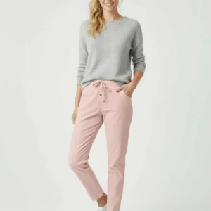 En kvinna står leende med ena handen i fickan. Hon bär en ljusgrå tröja, ljusrosa byxor med dragsko i midjan och vita sneakers. Hennes blonda hår är uppsatt i en lös hästsvans. Bakgrunden är enfärgad och ljus.