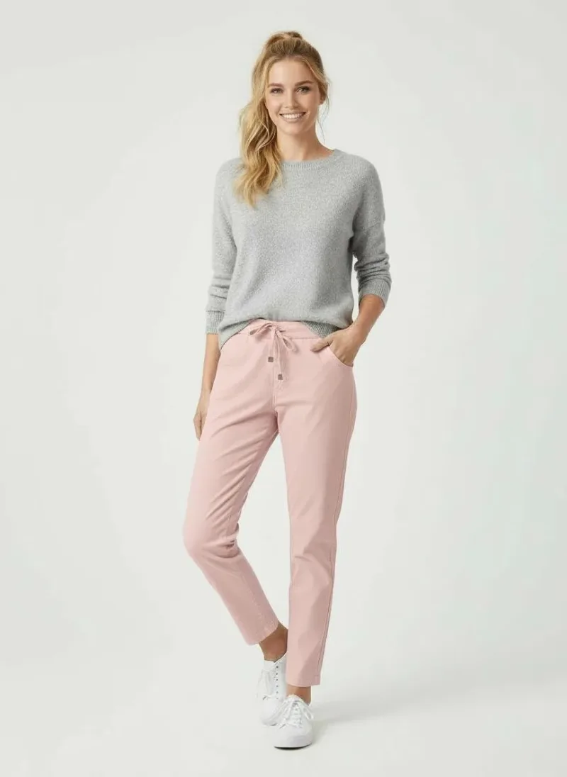 En kvinna står leende med ena handen i fickan. Hon bär en ljusgrå tröja, ljusrosa byxor med dragsko i midjan och vita sneakers. Hennes blonda hår är uppsatt i en lös hästsvans. Bakgrunden är enfärgad och ljus.