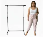 En kvinna i vitt linne och Cozy elastisk byxa i ljusrosa står barfota bredvid en tom klädställning i svart och silver mot en vit bakgrund.