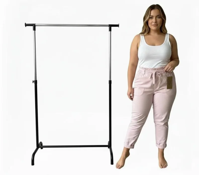 En kvinna i vitt linne och Cozy elastisk byxa i ljusrosa står barfota bredvid en tom klädställning i svart och silver mot en vit bakgrund.