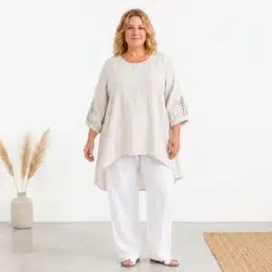 Woman wearing a loose beige linen tunic with crochet-sleeve details and white wide-leg pants in a bright studio setting.