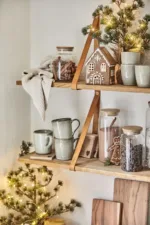 På två trähyllor står muggar, burkar med ingredienser och Pepparkakshus för ljus med snökristall. Små tallar i kruka med ljusslingor ger en festlig touch, medan skärbrädorna är staplade nedanför.