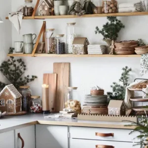 En mysig kökshörna dekorerad inför julen med Pepparkakshus för ljus med snökristall, trähyllor med tallrikar, muggar, burkar, vintergröna grenar, jultomtar och ett tänt ljus för en festlig stämning.