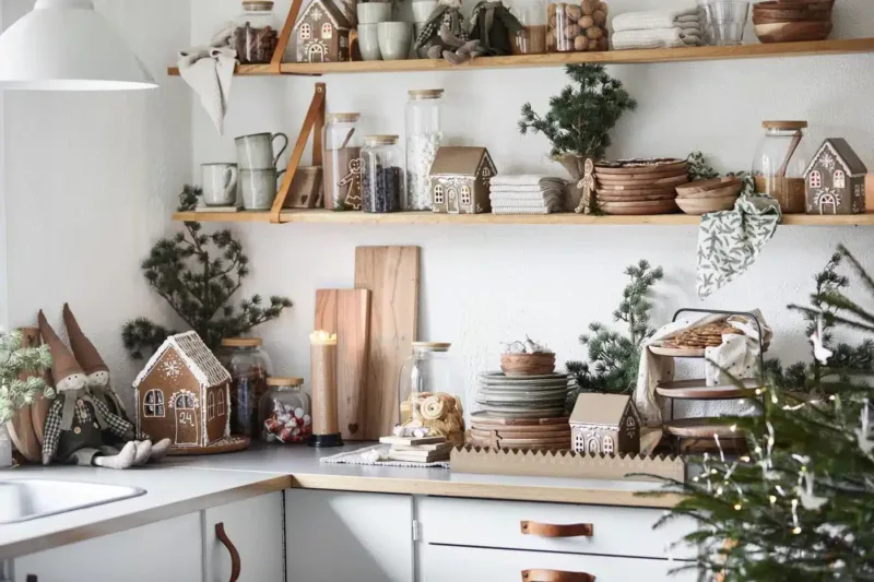 En mysig kökshörna dekorerad inför julen med Pepparkakshus för ljus med snökristall, trähyllor med tallrikar, muggar, burkar, vintergröna grenar, jultomtar och ett tänt ljus för en festlig stämning.
