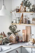 En mysig kökshörna julpyntad med Pepparkakshus för ljus med snökristall, godis i glasburkar, julmuggar, små krukväxter, skärbrädor och tygdockor på hyllor och bänkar.