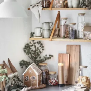 En mysig kökshörna julpyntad med Pepparkakshus för ljus med snökristall, godis i glasburkar, julmuggar, små krukväxter, skärbrädor och tygdockor på hyllor och bänkar.